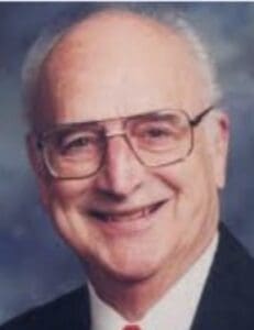 Elderly man smiling, wearing glasses and suit.
